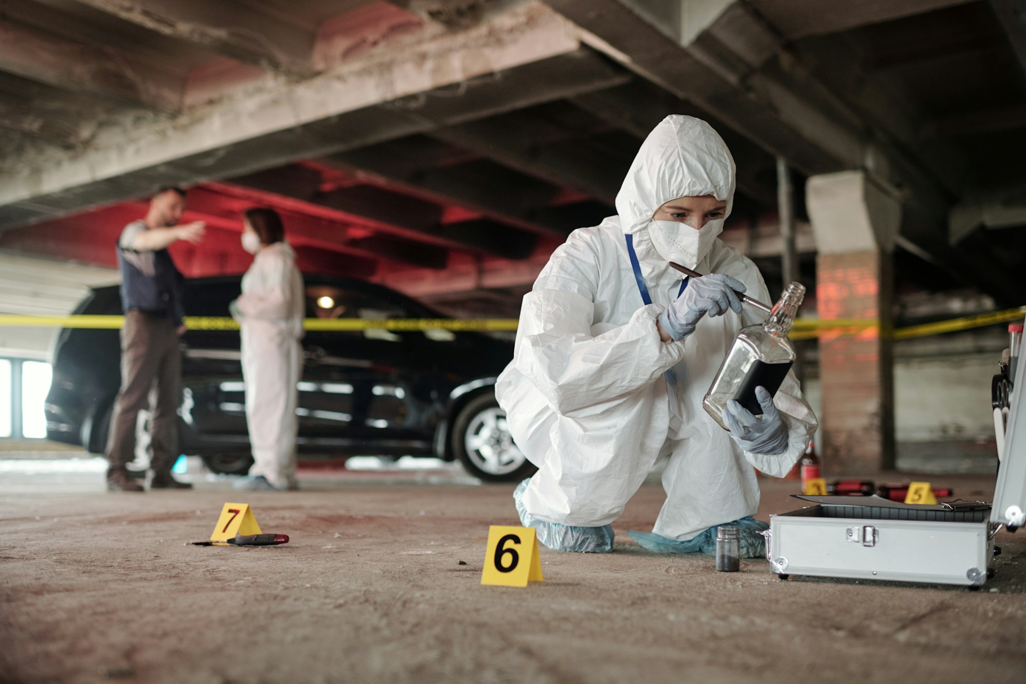 Young criminological expert in coveralls inspecting empty bottle on crime scene